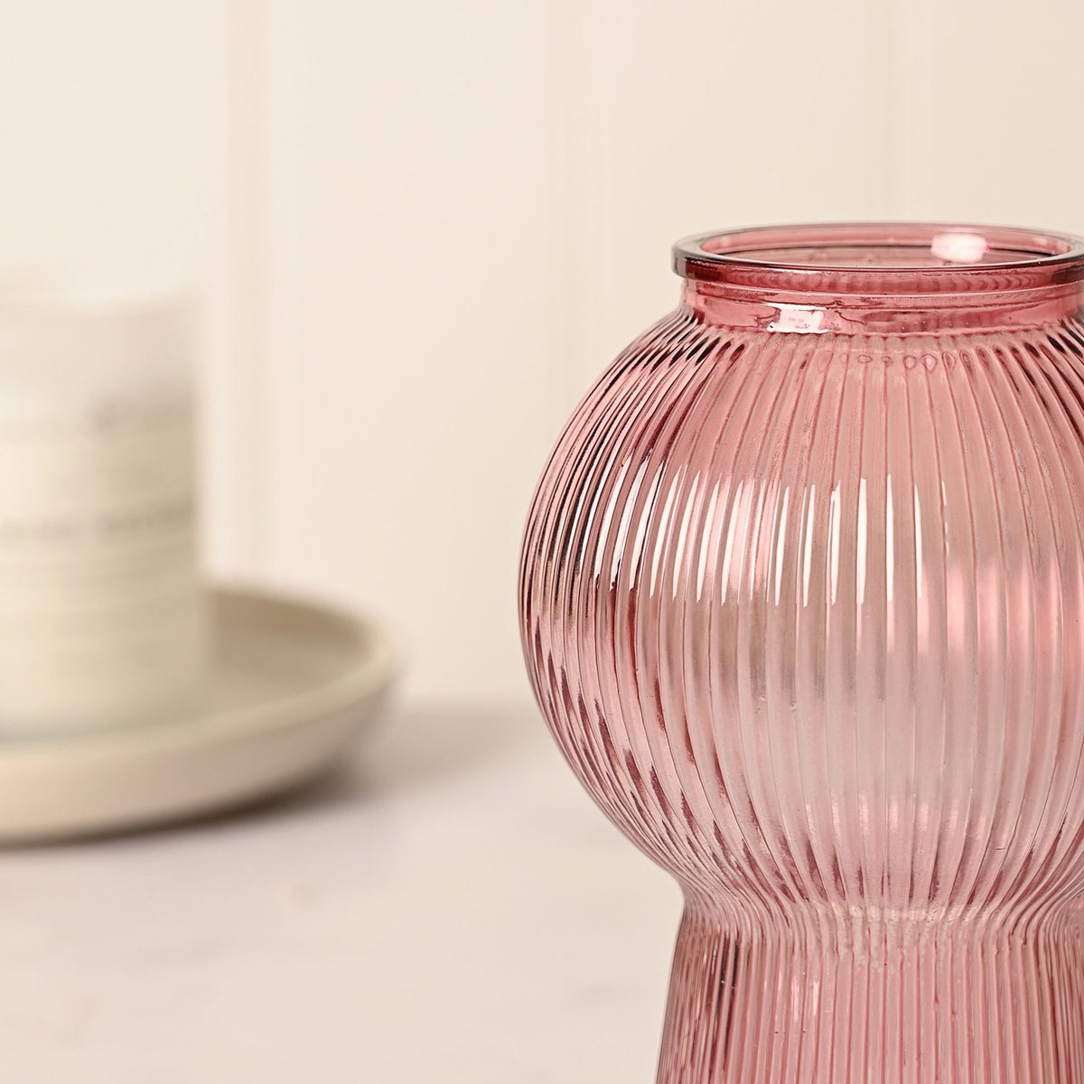 Ribbed Balloon Glass Vase with Flared Base in pink, close-up of the round ribbed body and neck, with a blurred ceramic jar and plate in the background.