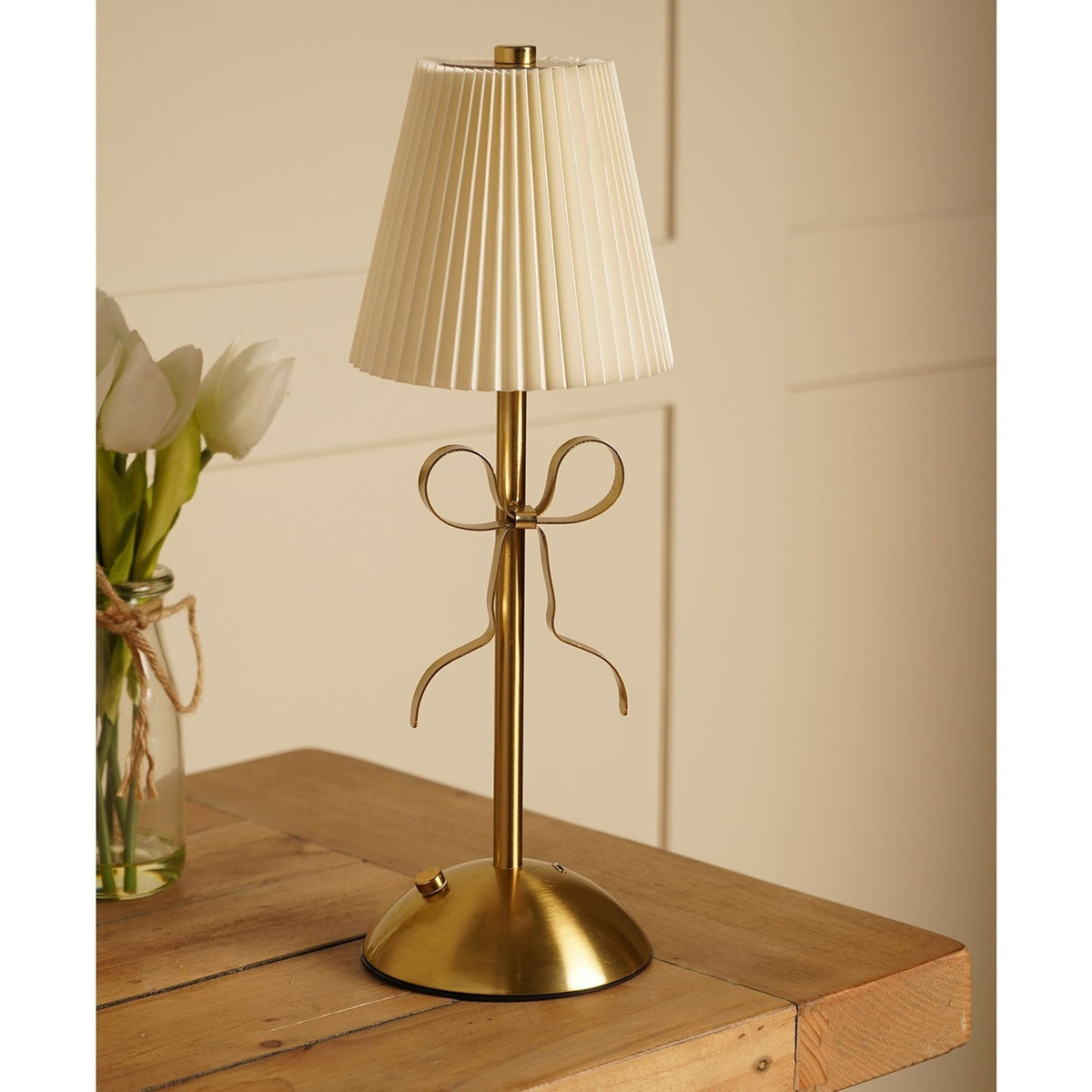 Brass Bow Pleated LED Touch Table Lamp with a cream pleated shade and gold ribbon bow detail on a slim stem, shown on a wooden table beside white tulips.