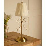 Brass Bow Pleated LED Touch Table Lamp with a cream pleated shade and gold ribbon bow detail on a slim stem, shown on a wooden table beside white tulips.