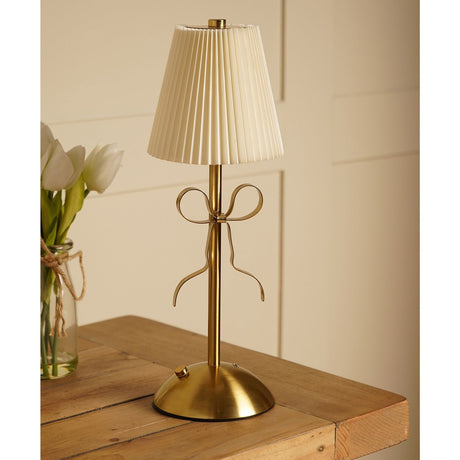 Brass Bow Pleated LED Touch Table Lamp with a cream pleated shade and gold ribbon bow detail on a slim stem, shown on a wooden table beside white tulips.