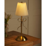 Brass Bow Pleated LED Touch Table Lamp with cream pleated shade and gold metal ribbon bow detail on stem, shown lit on a wooden table beside white tulips.