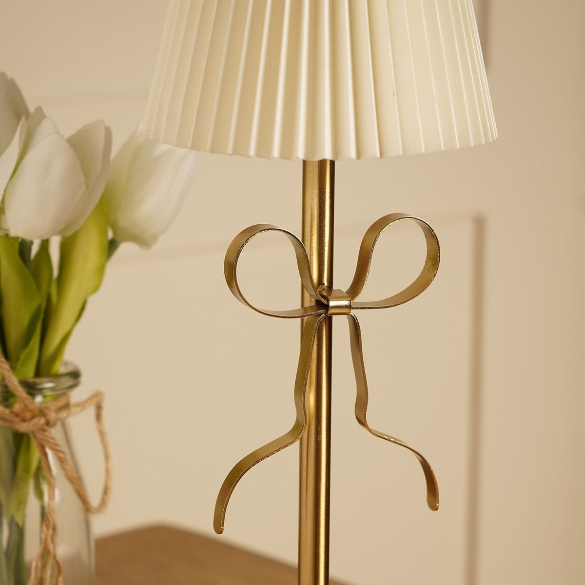 Close-up of the Brass Bow Pleated LED Touch Table Lamp's gold stem with a metal ribbon bow detail, beneath a cream pleated shade, near a vase of white tulips.