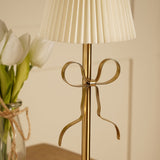 Close-up of the Brass Bow Pleated LED Touch Table Lamp's gold stem with a metal ribbon bow detail, beneath a cream pleated shade, near a vase of white tulips.