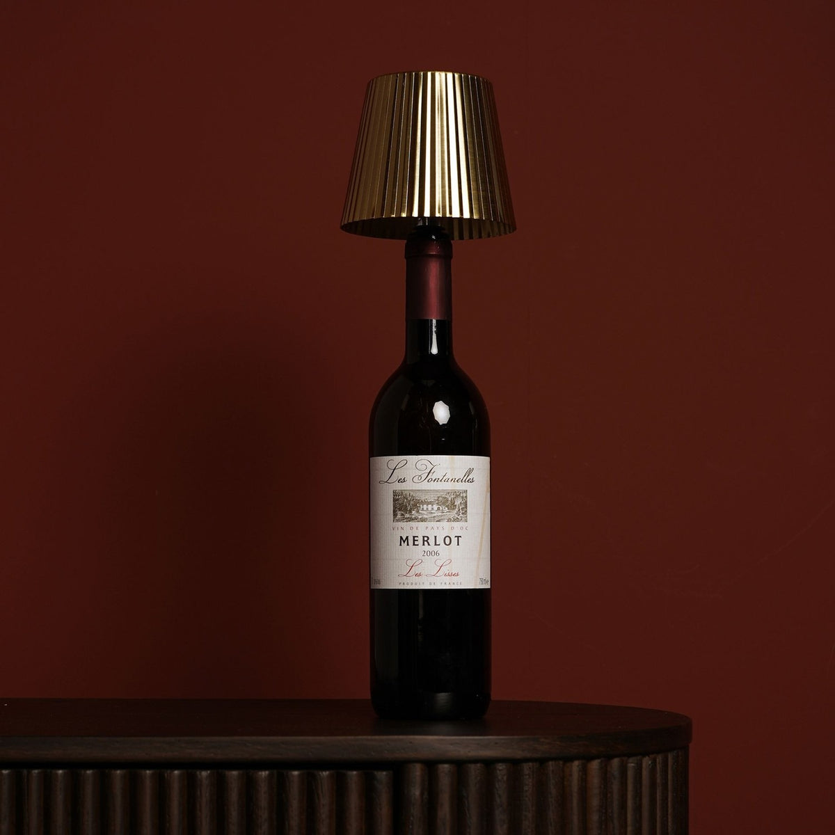 Wine Bottle Topper LED Touch Metal Table Lamp with gold pleated tapered shade and white touch top, fitted onto a Merlot bottle on a dark sideboard against a deep red background.