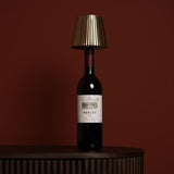 Wine Bottle Topper LED Touch Metal Table Lamp with gold pleated tapered shade and white touch top, fitted onto a Merlot bottle on a dark sideboard against a deep red background.
