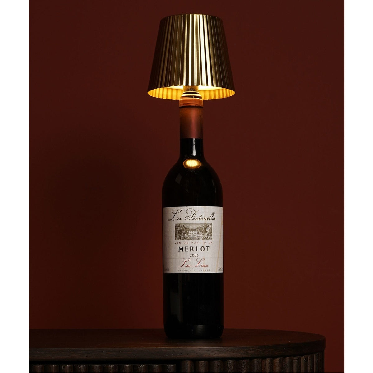 Wine bottle topper LED touch table lamp with a gold pleated tapered shade and white top, fitted onto a Merlot bottle on a dark wood table against a maroon background.