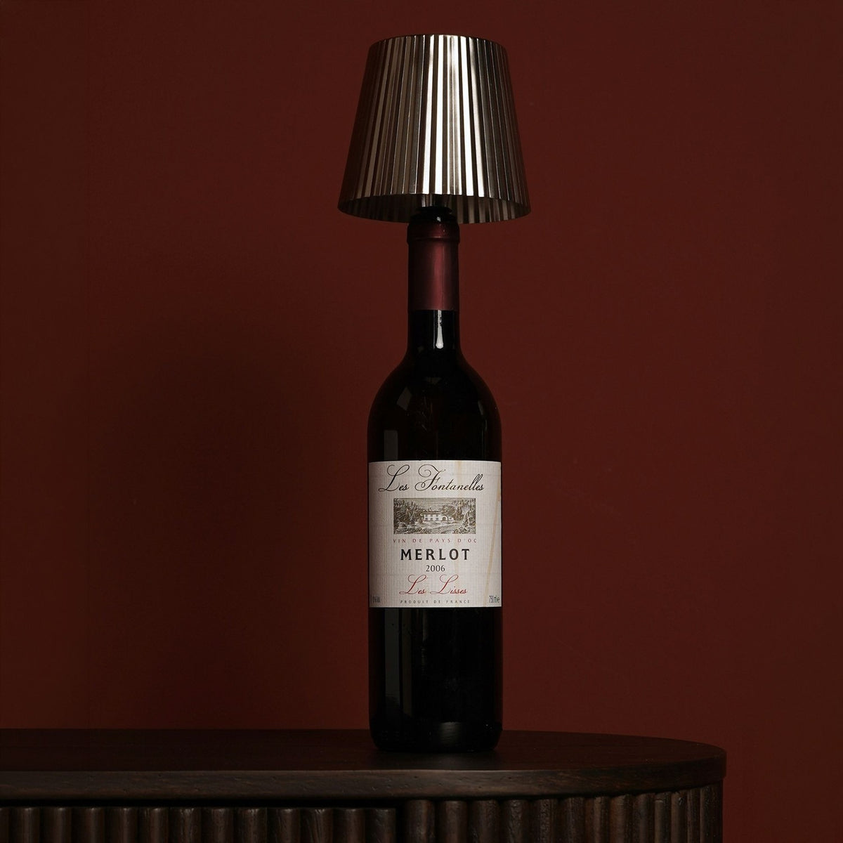Round Wine Bottle Topper LED table lamp in silver metal with a pleated tapered shade, fitted onto a Merlot wine bottle against a dark red background.