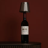 Round Wine Bottle Topper LED table lamp in silver metal with a pleated tapered shade, fitted onto a Merlot wine bottle against a dark red background.