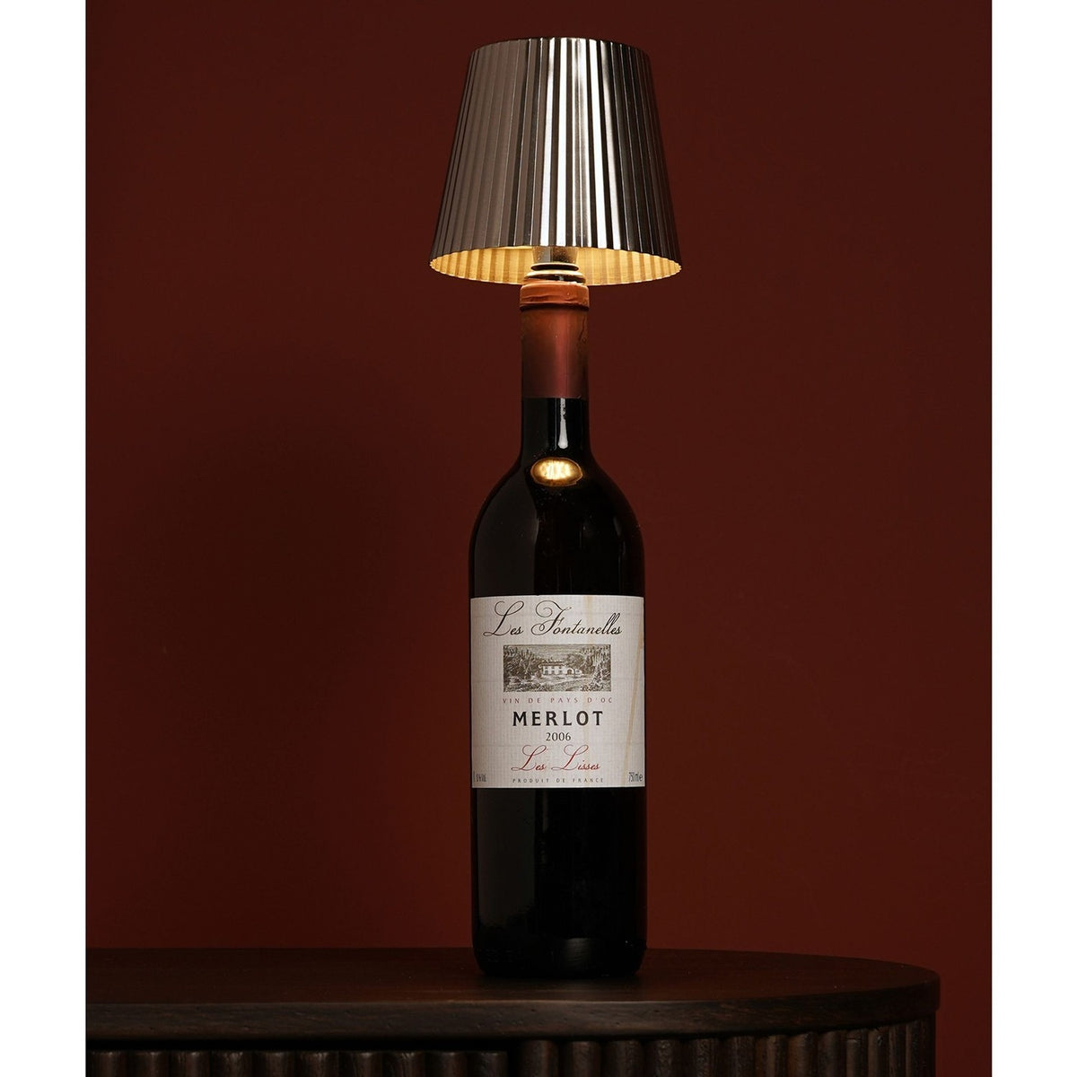 Round Wine Bottle Topper LED touch table lamp in silver metal with a pleated tapered shade, fitted into a Merlot wine bottle on a dark wood table against a maroon background.