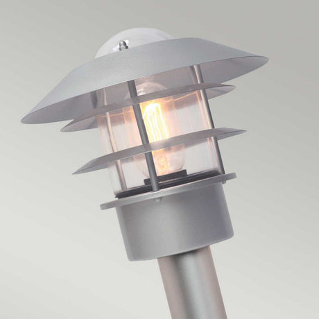 Close-up of the Helsingor 1 Light Bollard Lantern in silver, showcasing its modern, sleek metallic design that evokes Scandinavian aesthetics. This lantern features a radiant bulb enclosed in a transparent casing with layered shades, set against a light gray background.