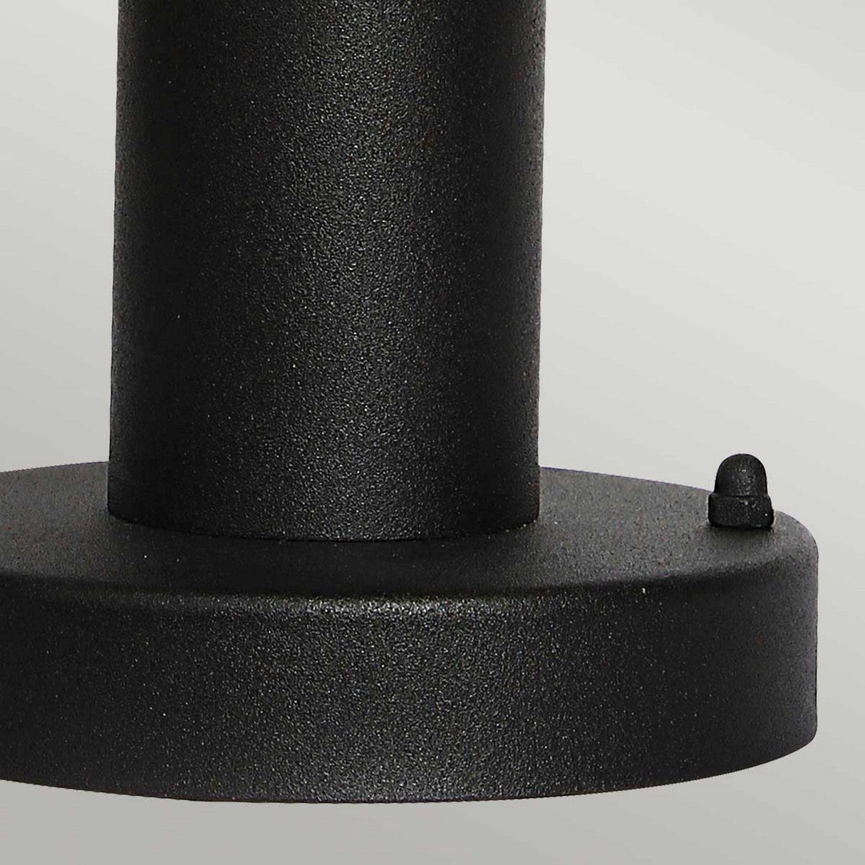 Close-up of the Helsingor 1 Light Pedestal Light - Black, featuring a black metal cylindrical shape attached to a flat circular base, capturing the elegant simplicity of Scandinavian design. A small round knob adorns the edge of the base, evoking the look of an outdoor lighting fixture against a plain light gray background.