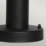 Close-up of the Helsingor 1 Light Pedestal Light - Black, featuring a black metal cylindrical shape attached to a flat circular base, capturing the elegant simplicity of Scandinavian design. A small round knob adorns the edge of the base, evoking the look of an outdoor lighting fixture against a plain light gray background.
