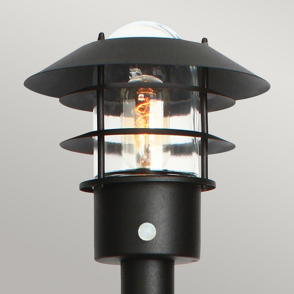 The Helsingor 1 Light Bollard Lantern with PIR in black is a modern outdoor lamp with an IP44 rating and a sturdy metal frame. It features a clear glass enclosure that reveals the glowing bulb inside, set against a plain gray background, highlighting its sleek contemporary design.