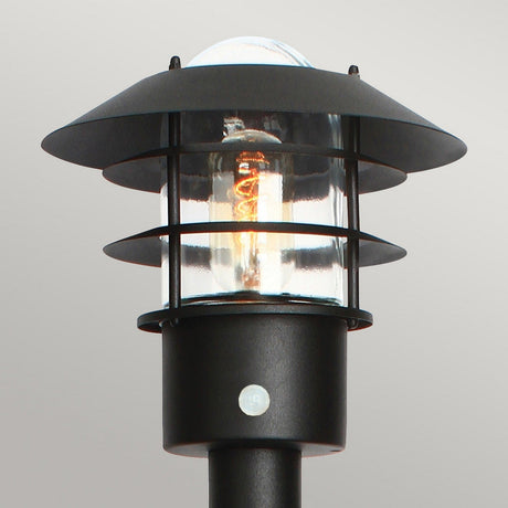 The Helsingor 1 Light Bollard Lantern with PIR in black is a modern outdoor lamp with an IP44 rating and a sturdy metal frame. It features a clear glass enclosure that reveals the glowing bulb inside, set against a plain gray background, highlighting its sleek contemporary design.