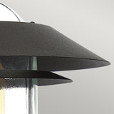 A close-up of the Helsingor 1 Light PIR Pedestal Light showcases its modern design with an angular metallic shade. The fixture, boasting a sleek and contemporary look, comes in a black finish with a clear glass bulb cover, making it perfect for security lighting. The background is a plain light gray.