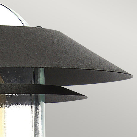 A close-up of the Helsingor 1 Light PIR Pedestal Light showcases its modern design with an angular metallic shade. The fixture, boasting a sleek and contemporary look, comes in a black finish with a clear glass bulb cover, making it perfect for security lighting. The background is a plain light gray.