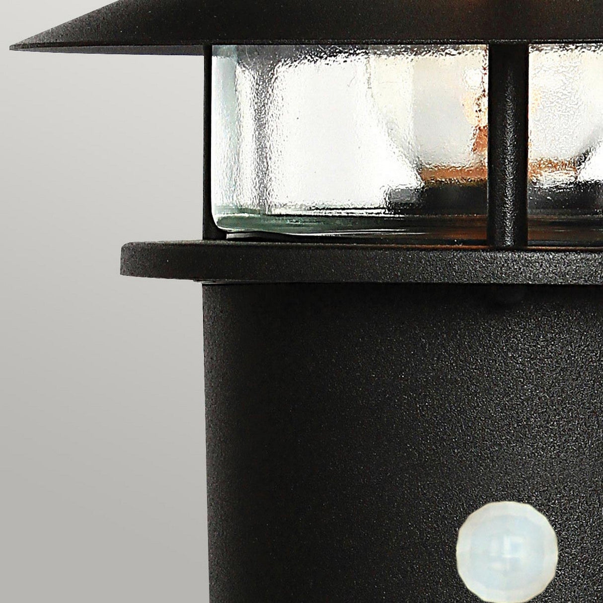 Close-up of the Helsingor 1 Light PIR Pedestal Light in black, featuring a glass enclosure and partially visible against a plain gray background. The lantern showcases a round, textured base with a visible screw and emits a warm glow, ideal for providing security lighting on any evening.