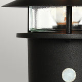 Close-up of the Helsingor 1 Light PIR Pedestal Light in black, featuring a glass enclosure and partially visible against a plain gray background. The lantern showcases a round, textured base with a visible screw and emits a warm glow, ideal for providing security lighting on any evening.