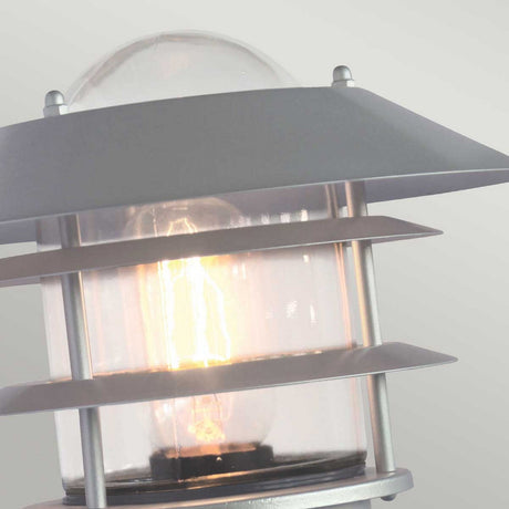A close-up of the Helsingor 1 Light Outdoor PIR Wall Light in silver shows its energy-efficient design, featuring a clear glass casing, metal accents, and a visible glowing bulb inside. This sleek, industrial fixture enhances security lighting against a plain background.