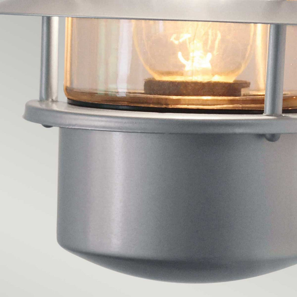 A close-up of the Helsingor 1 Light Outdoor Wall Light in silver, featuring a stainless steel industrial-style design with a clear glass cover that reveals the glowing bulb inside, set against a soft, light gray background.