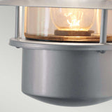 A close-up of the Helsingor 1 Light Outdoor Wall Light in silver, featuring a stainless steel industrial-style design with a clear glass cover that reveals the glowing bulb inside, set against a soft, light gray background.