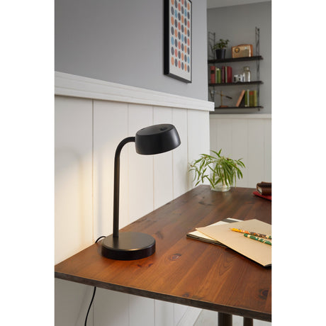 Hemarne LED Table Lamp in black on a wooden desk, with a round shallow head on a flexible gooseneck arm and round base, shown lit in a home office setting.