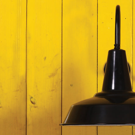 Hex Industrial Factory Swan Neck Wall Light in black, with a wide shallow shade and tapered top on a curved arm, mounted against a yellow wooden panel wall.