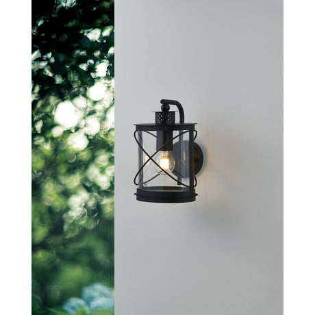 Hilburn black outdoor wall light with lantern-style cylindrical shade and crossed hoop wires, mounted on a light grey exterior wall near greenery.