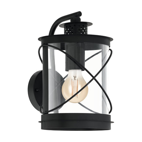 Black Hilburn outdoor wall light with lantern-style design, hooked mounting arm, clear cylindrical shade, crossed hoop wires, perforated top cap, and visible E27 bulb.