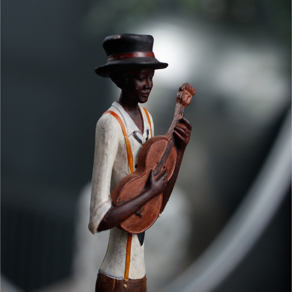 Hill Interiors Nerisse figurine of a jazz musician in a black hat, white shirt with orange braces, playing a wooden guitar, shown against a blurred background.