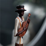 Hill Interiors Nerisse figurine of a jazz musician in a black hat, white shirt with orange braces, playing a wooden guitar, shown against a blurred background.