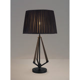The Hilton 1 Light Small Wood Table Lamp features a black and grey geometric base with a pleated black shade, casting a soft glow as it sits on a light surface against a plain grey background.