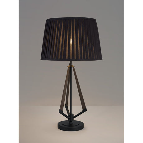 The Hilton 1 Light Small Wood Table Lamp features a black and grey geometric base with a pleated black shade, casting a soft glow as it sits on a light surface against a plain grey background.