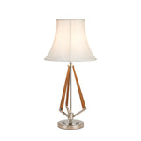 The Hilton 1 Light Wood Table Lamp features a nickel and taupe finish, cream shade, wooden angled supports, and a round chrome base, shown against a plain white background.