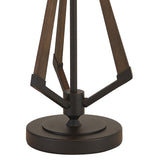 A close-up of the Hilton 1 Light Wood Table Lamp shows its round, dark metal base with angled wooden supports in black and grey, joined by metal brackets, topped with a taupe/halo gold shade.
