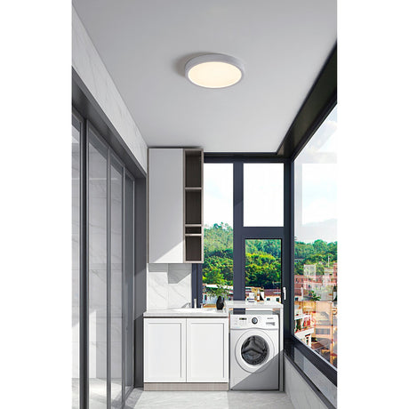 Hinanne 28.5cm round LED downlight in white steel with a flush white plastic diffuser, shown fitted to the ceiling of a bright laundry room with floor-to-ceiling windows.