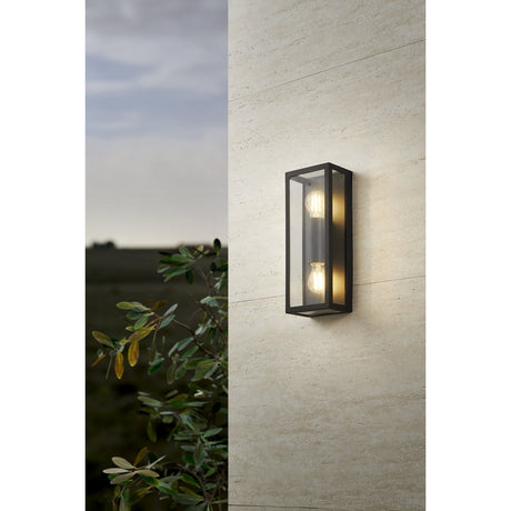 Hirbessa 2 Light Outdoor Wall Light in black steel with clear glass panels, two visible E27 bulbs illuminated, mounted on a beige stone wall beside a plant.