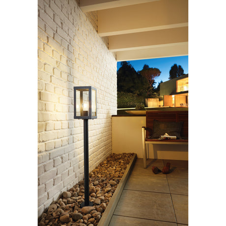 Hirbessa Bollard Light 101.5cm in black, square glass-panelled lantern head on a slim post, illuminating a brick wall beside a pebble border on an outdoor patio.