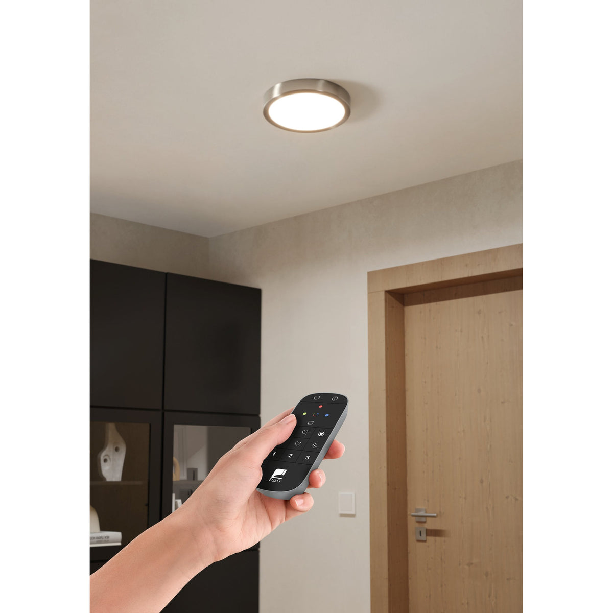 Hand holding the Hismena Zigbee Remote Control in grey, a slim handheld lighting controller with black button face, shown in a hallway with ceiling light and wooden door visible.