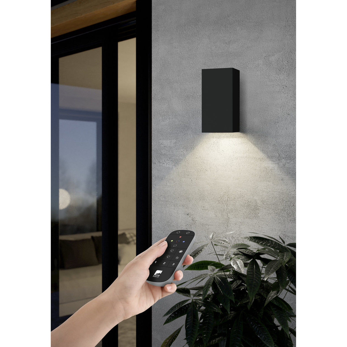 Hand holding the Hismena Zigbee Remote Control in grey with black button face, used to operate a black rectangular up-down wall light outdoors near a glass door and plant.