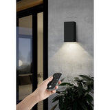 Hand holding the Hismena Zigbee Remote Control in grey with black button face, used to operate a black rectangular up-down wall light outdoors near a glass door and plant.