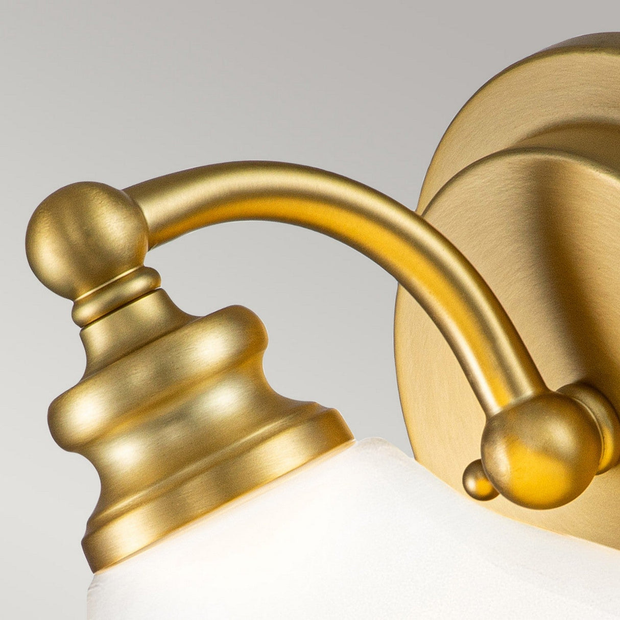 A close-up of the Ainsley 1 Light Wall Light in a brushed brass finish showcases its elegant wall-mounted design with a curved arm and an opal glass shade. The vintage style is enhanced by the energy-efficient LED lamp, providing sustainable illumination.
