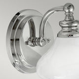 The Ainsley 1 Light Wall Light, showcasing a polished chrome finish, is a contemporary wall-mounted fixture with a round base. It features a curved arm that holds an opal glass shade, creating a striking contrast against the plain gray background with its sleek design.