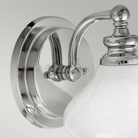 The Ainsley 1 Light Wall Light, showcasing a polished chrome finish, is a contemporary wall-mounted fixture with a round base. It features a curved arm that holds an opal glass shade, creating a striking contrast against the plain gray background with its sleek design.