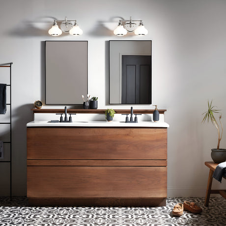 A modern bathroom showcases a wooden vanity with a white countertop paired with twin black faucets. Above the vanity, two rectangular mirrors are complemented by polished chrome Ainsley 2 Light Wall Lights featuring LED lamps. The space is adorned with a small plant and toiletries, while patterned tiles cover the floor.