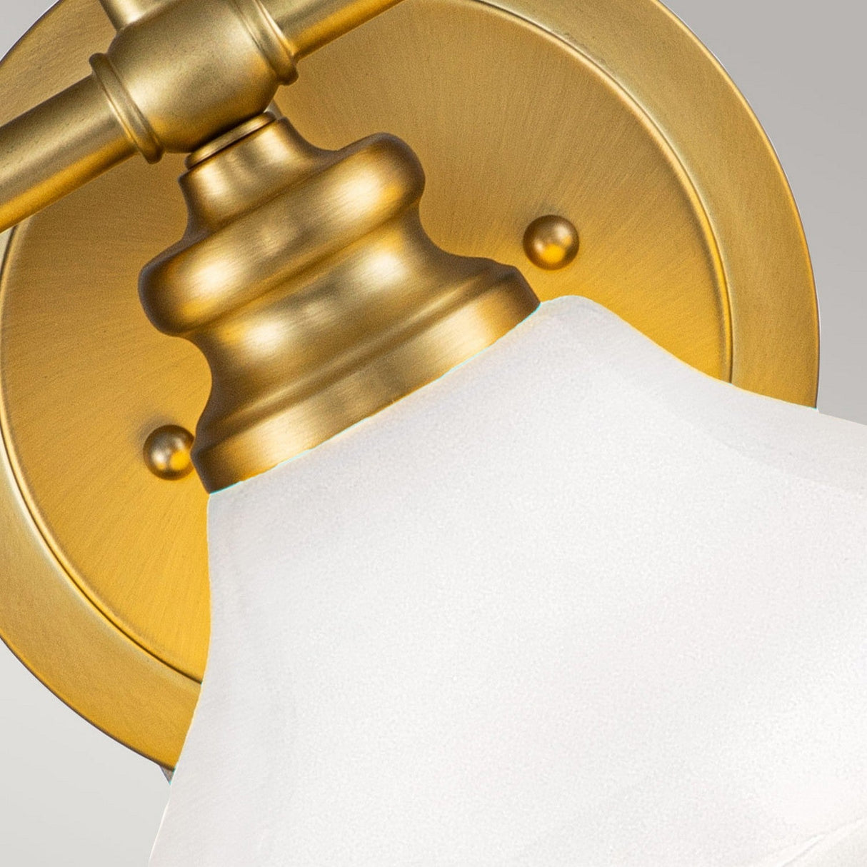 Close-up of the Ainsley 3 Light Wall Light in brushed brass, showcasing its gold wall-mounted design with an opal glass shade. This fixture features a round base attached to the wall, and a graceful curve supporting the shade, providing energy-efficient LED illumination against a plain gray background.