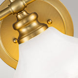 Close-up of the Ainsley 3 Light Wall Light in brushed brass, showcasing its gold wall-mounted design with an opal glass shade. This fixture features a round base attached to the wall, and a graceful curve supporting the shade, providing energy-efficient LED illumination against a plain gray background.