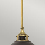 A close-up of the Amelia 1 Light Mini Pendant - Bronze showcases an elegant light fixture with a brass rod and dark metallic shade. Enhanced by its opal glass dome, this bronze lighting piece stands out beautifully against a light gray background.