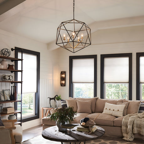 The modern living room boasts a beige sectional sofa along with an Astrid 5 Light Chandelier in Matte Bronze & Gold, which casts a warm glow throughout the space. A round coffee table enhances the overall style, while shelves are adorned with decorative items. Large windows fitted with shades allow natural light to flood the room, illuminating its neutral tones and creating a cozy atmosphere.