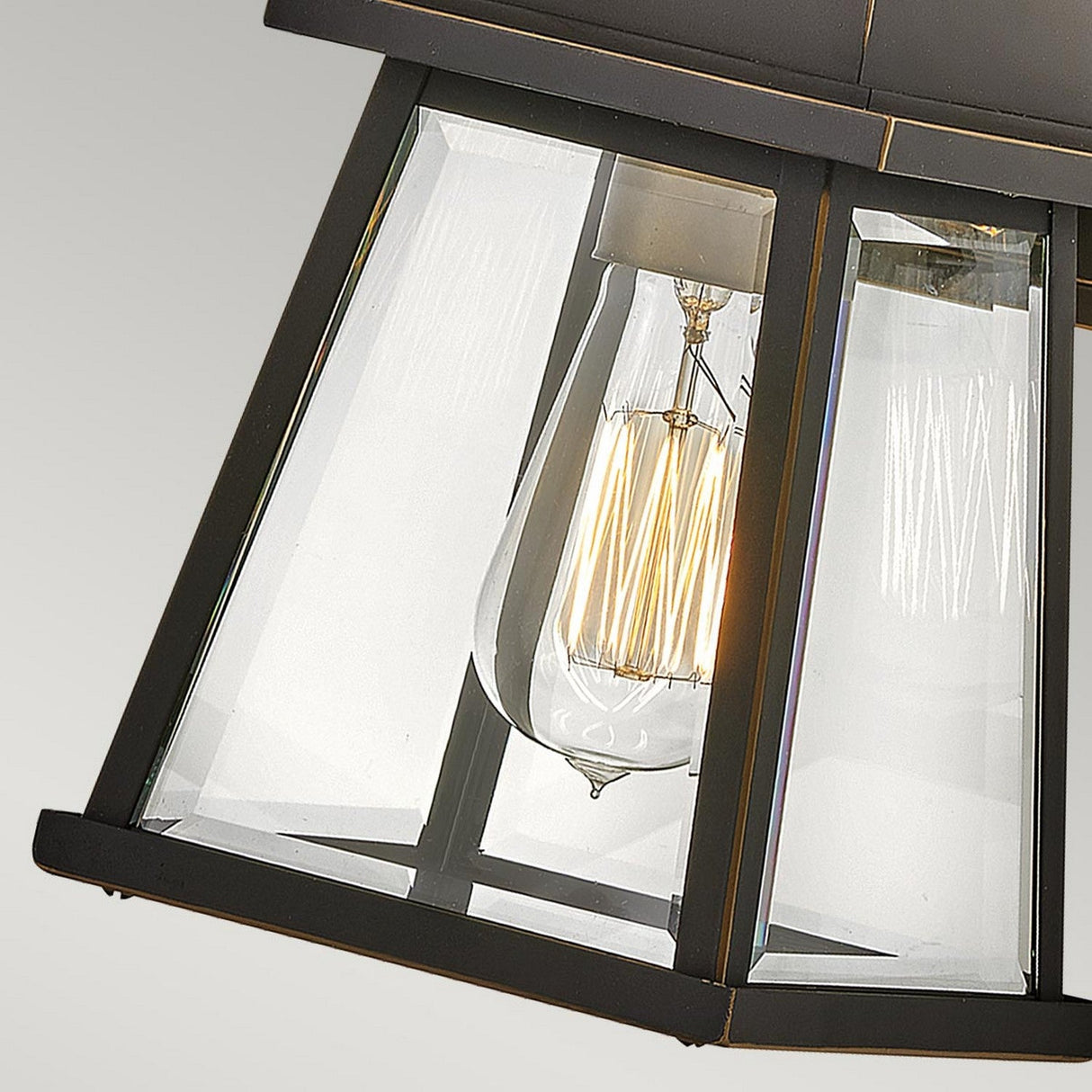 Close-up of the Bainbridge 1 Light Outdoor Small Wall Lantern in a modern bronze finish, featuring bevelled glass panels and showcasing an illuminated vintage-style filament bulb. The background is a neutral gray, which highlights the intricate details and design of the lantern.
